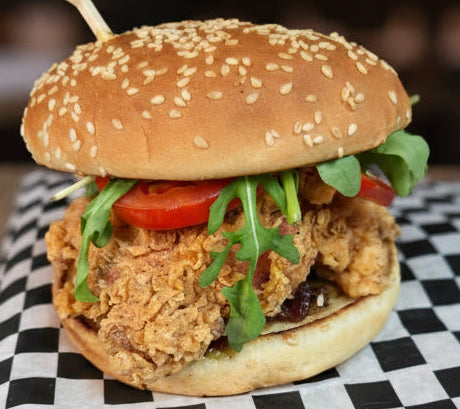 A close-up of a gourmet fried chicken burger with crispy golden-brown fried pieces, fresh green arugula, red tomato slices, and a sesame seed bun, resting on a black and white checkered paper liner.