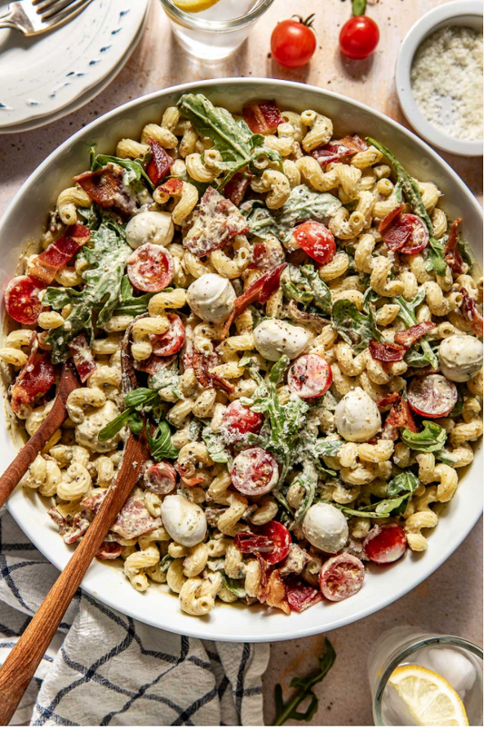A large white bowl filled with a pasta salad containing penne pasta, mozzarella balls, sliced tomatoes, greens, and bacon bits, served with wooden serving utensils on a table setting.