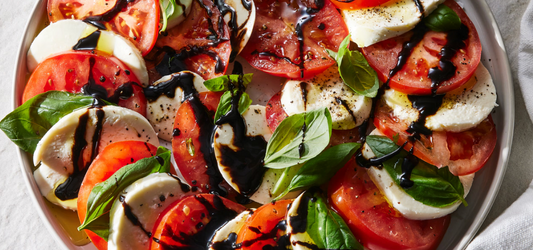 A close-up overhead view of a Caprese salad in a white bowl, featuring sliced red tomatoes, white mozzarella cheese, fresh green basil leaves, and a drizzle of dark balsamic glaze.
