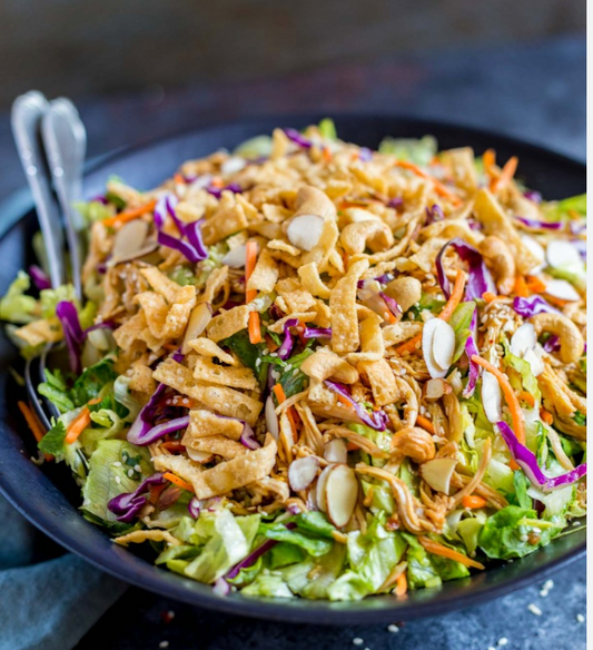 A close-up shot of a fresh salad in a black bowl, featuring shredded lettuce, carrots, purple cabbage, crispy fried strips, and sliced almonds, with two silver forks on the left.